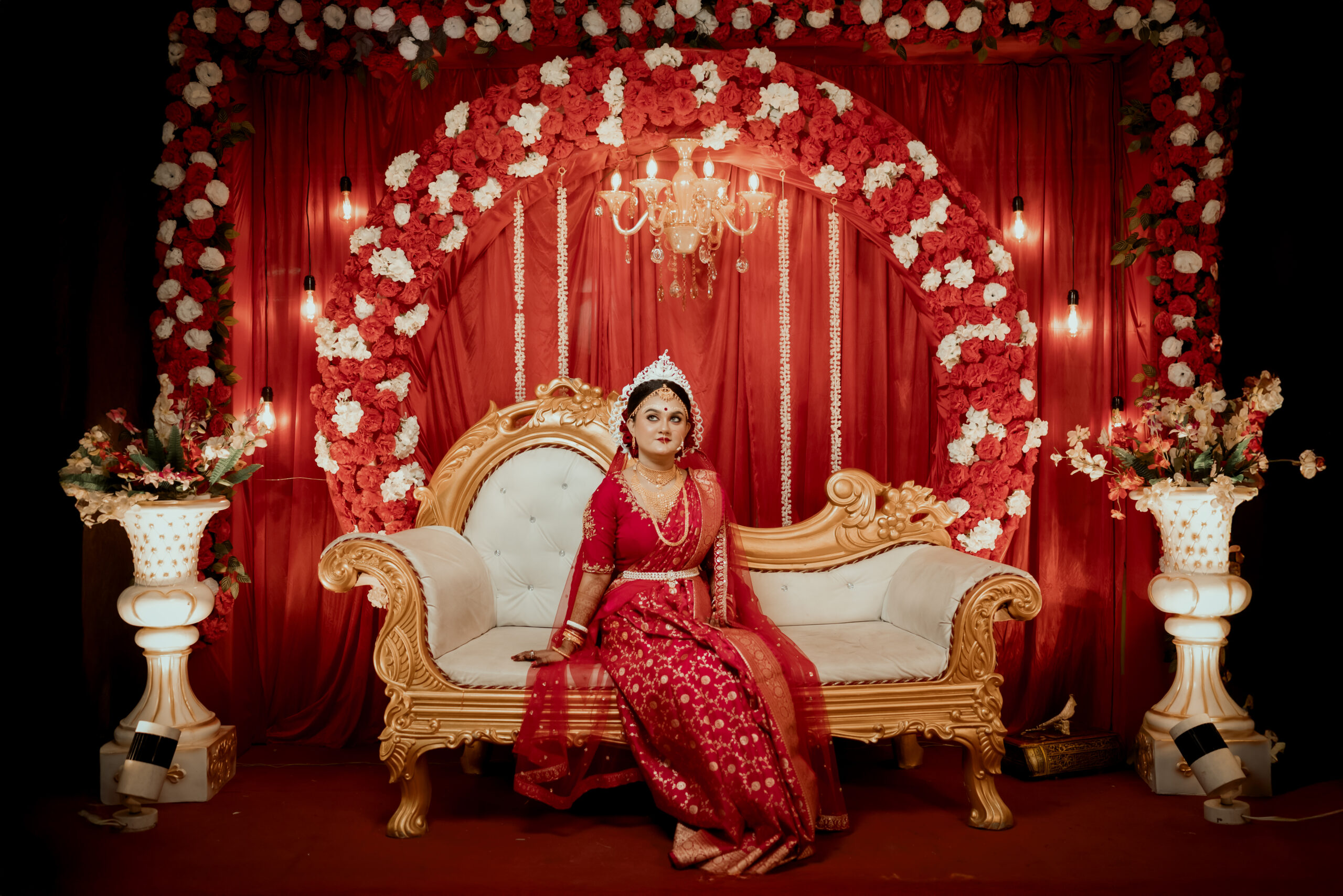 Bride in red wedding attire seated beneath an ornate red backdrop
