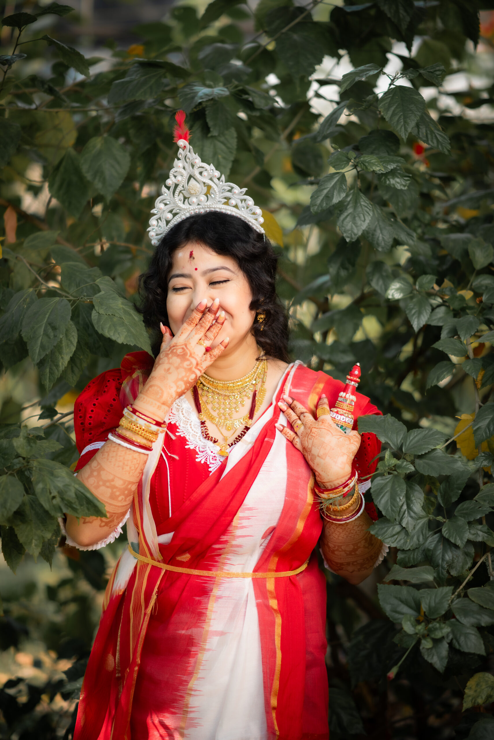Bengali bride laughing in a red and white sari beneath green foliage